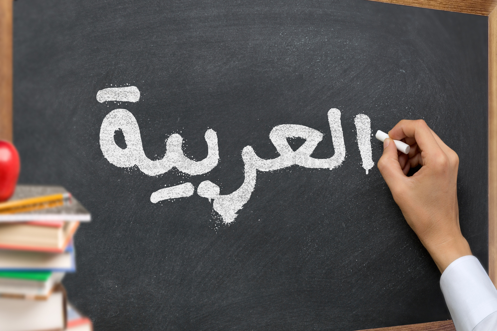 Hand writing on a blackboard in a Arabic language learning class course with the text "Arabic" written on it. with Some books and school materials concept.